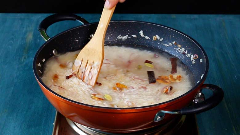 A slotted wooden spatula is seen stirring the rice topped with water before bringing it to a boil in an orange non stick pan on an electric stove on a blue wooden countertop - Kerala Ghee Rice with California Walnuts Recipe